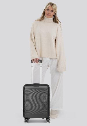 Valise rigide noire avec un motif en vagues, une poignée télescopique et quatre roues. Entourée d'une personne portant un pull crème et un pantalon blanc.