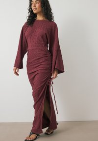 Losse bordeauxrode jurk van textuurstof, met lange klokmouwen, gerimpelde details en een zijsplit dat met een koord om de taille is vastgebonden.