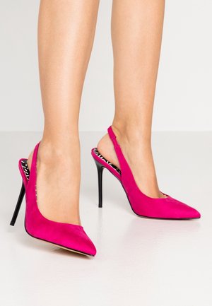 Fødder iført klare pink spidse slingback-højhælede sko med sorte stiletter på en glat hvid overflade.