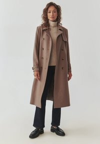 Brauner Wollmischung-Trenchcoat mit doppelreihiger Front, ärmellosem Kragen, seitlichen Taschen und Ärmeln. Getragen über einem cremefarbenen Rollkragenpullover und dunklen Hosen.