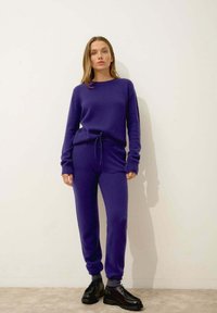 Lila stickad lounge-set som består av en långärmad topp med rund hals och matchande byxor med dragsko. Svarta snörskor fullbordar outfiten.