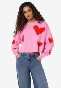 Ροζ κολαριστό πουλόβερ με πολλαπλά κόκκινα σχέδια καρδιάς, με μακριά μανίκια και στρογγυλή λαιμόκοψη, συνδυασμένο με τζιν παντελόνια ψηλής μέσης.