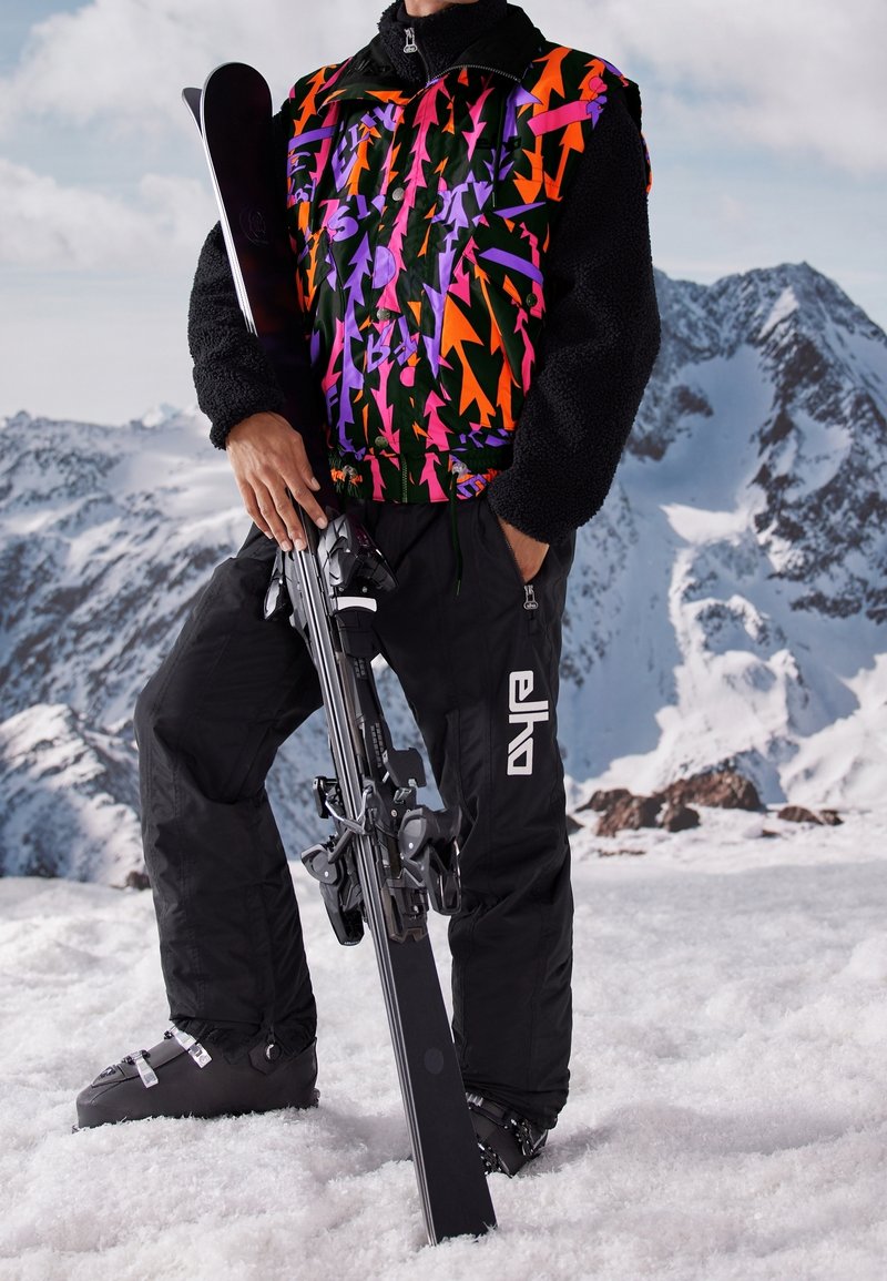 Person in farbiger Skijacke und schwarzen Hosen hält Skier und steht auf einem verschneiten Berg mit verschneiten Gipfeln im Hintergrund.