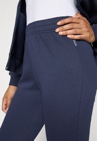 Personne portant un pantalon de survêtement Champion bleu marine et une veste assortie, avec une main reposant sur la taille.
