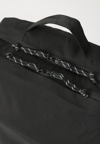 Sac à dos noir doté de deux zips supérieurs avec des cordons de tirage rayés noir et blanc et une solide poignée de transport. Tissu lisse et texturé.
