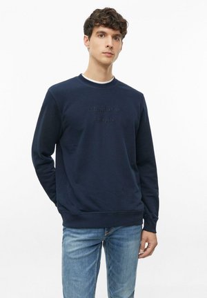 Junger Mann trägt ein marineblaues Sweatshirt mit dem Text "all I need is time and space" und blaue Jeans, steht mit einer Hand hinter dem Rücken.