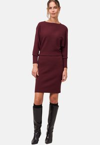 Robe tricotée bourgogne avec un haut oversized et une jupe ajustée. Associée à des bottes noires en cuir verni jusqu'au genou et des chaussettes noires ajustées.