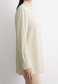 Camicia button-up beige chiaro con colletto, maniche lunghe e vestibilità morbida. Tessuto liscio con leggere arricciature sulle maniche e cuciture laterali.