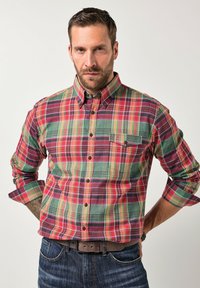 Uomo con barba e capelli corti che indossa una camicia a quadri multicolore con le maniche arrotolate, jeans scuri, cintura marrone, in piedi con le mani dietro la schiena.