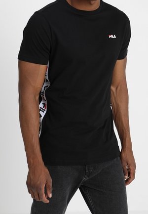 Man in zwarte crewneck FILA t-shirt met een witte en rode logo-streep langs de zijkanten en donkere jeans, staand tegen een witte achtergrond.