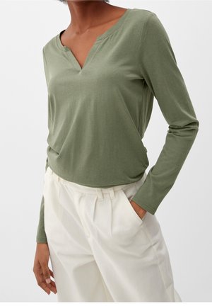 Osoba ubrana w oliwkowozieloną koszulę z długim rękawem i dekoltem w serek, wpuszczoną w wysokie, luźne białe spodnie, z jedną ręką w kieszeni.