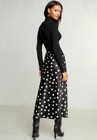 Schwarzer gerippter Rollkragenpullover kombiniert mit einem langen, schwarzen Satinrock mit großen weißen Punkten. Das Outfit beinhaltet kniehohe schwarze Stiefel.
