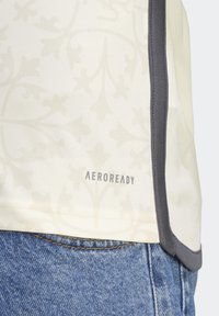 Camisa de un suave marfil, confeccionada con una tela de sutil patrón floral, con ribetes grises y un logo "AEROREADY" visible en el dobladillo.
