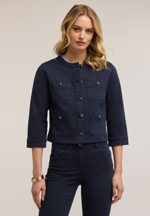 Vrouw met blond haar die een donkerblauw jasje met knopen draagt, met vier zakken aan de voorkant, en bijpassende broek, staand tegen een effen achtergrond.