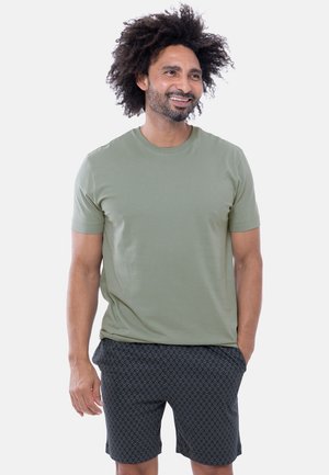 Man met krullend haar die een lichtgroen t-shirt en donkere korte broek met patroon draagt, glimlachend en staand met één hand in de zak.