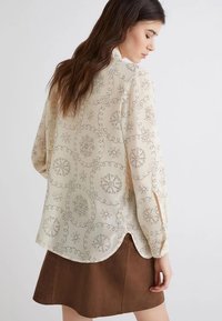 Blusa color crema con motivi floreali e circolari, maniche lunghe e orlo arrotondato. Il tessuto appare leggero e liscio.
