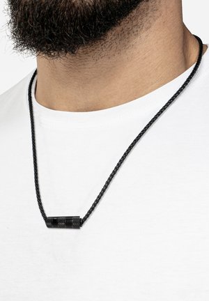Black braided necklace featuring a cylindrical metal pendant with textured accents, worn over a white t-shirt with a round neckline.