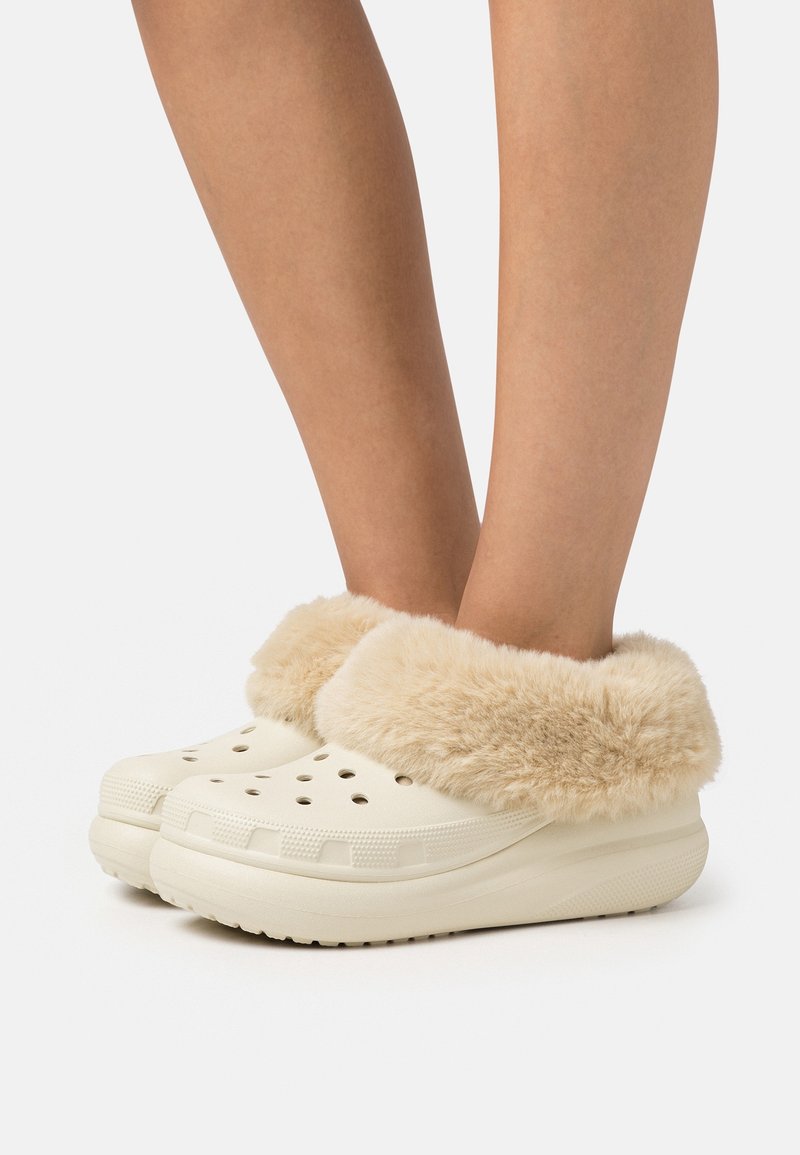 Pieds portant des sabots crème à enfiler avec des trous de ventilation et une épaisse doublure en fausse fourrure beige autour des chevilles.