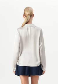 Vrouw met een blonde paardenstaart, gekleed in een lichtbeige blouse met lange mouwen en een marineblauwe geplooide rok, gezien van achteren tegen een effen witte achtergrond.