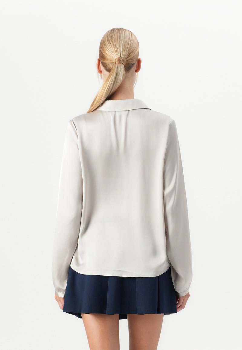 Femme avec une queue de cheval blonde portant un blouse à manches longues beige clair et une jupe plissée bleu marine, vue de dos sur fond blanc uni.