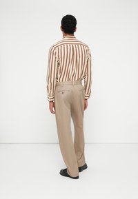 Ριγέ καφέ και κρεμ πουκάμισο με μακριά μανίκια, συνδυασμένο με ανοιχτό μπεζ παντελόνι. Έχει πίσω τσέπες και μαύρα παπούτσια.