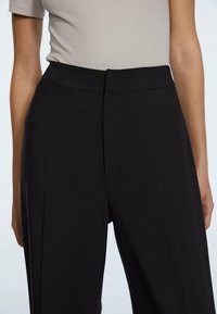 Black high-waisted trousers with a smooth fabric texture, featuring a flat front, pleats, and a tailored design.