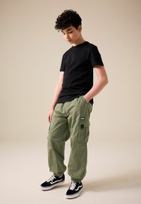 T-shirt nera a maniche corte abbinata a pantaloni cargo leggeri verde oliva con tasche e polsini elasticizzati, completata da sneakers nere e bianche.