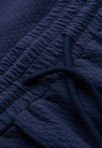 Gros plan d’un tissu texturé bleu marine avec taille élastique et cordon noué, probablement une partie d’un pantalon ou short décontracté.