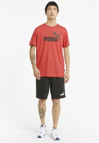 Červené tričko s krátkým rukávem a černým logem Puma, doplněné černými šortkami s bílým logem Puma, nošené s bílými teniskami.