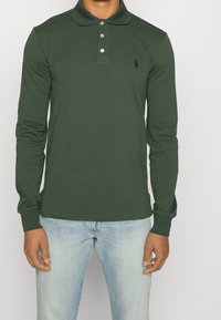 Tmavě zelená polo košile s dlouhým rukávem vyrobená z bavlny, s límečkem, tříbuttonovou légou a černým logem na hrudi.