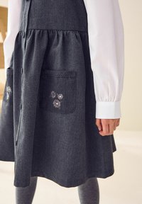 Donkergrijze denim jurk met een uitlopende rok, voorzien van twee voorzakken versierd met bloemenborduursels. Witte blouse met lange mouwen eronder.