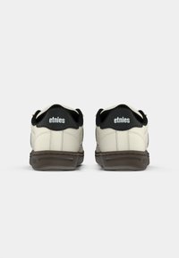 Sapatilhas de cor creme com detalhes em preto, apresentando um cabedal em couro liso, colarinho acolchoado e uma sola de borracha texturizada em castanho. Logotipo da Etnies visível.