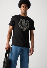 Camiseta negra con un gráfico de la cara de un tigre en líneas blancas. Combinada con jeans de mezclilla azules y una bolsa de mensajero negra.