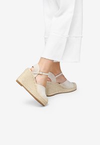 Sandales compensées beiges avec une tige en tissu et une semelle en jute tressé. Présentent une bride au niveau de la cheville et un talon ouvert, posées sur un pantalon blanc.