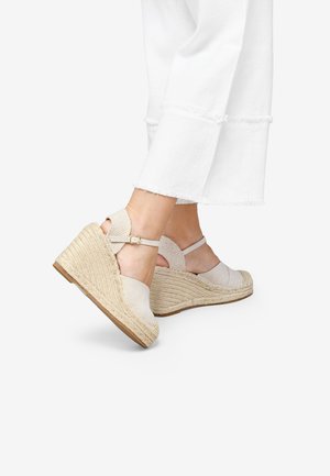 Sandali con zeppa beige con tomaia in tessuto e suola in juta intrecciata. Presentano una cinghia alla caviglia e un tallone aperto, posizionati contro pantaloni bianchi.