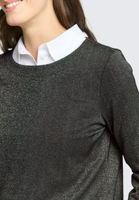 Czarna, błyszcząca koszula z długim rękawem i okrągłym dekoltem, noszona na białej koszuli z kołnierzykiem pod spodem. Gładka faktura, minimalistyczne detale.