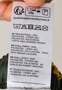 Waslabel met details over de stofinhoud: 100% polyester buitenkant, 90% polyester en 10% elastaan voering. Verzorgingsinstructies aangegeven met symbolen.