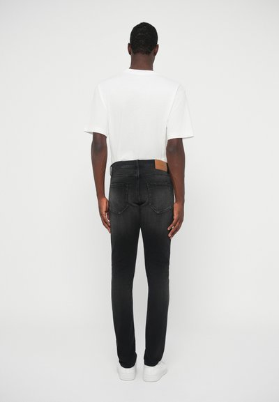 Άνδρας που φοράει λευκό μπλουζάκι, ξεθωριασμένα μαύρα τζιν και λευκά αθλητικά παπούτσια, στέκεται με την πλάτη γυρισμένη σε έναν απλό φωτεινό φόντο.