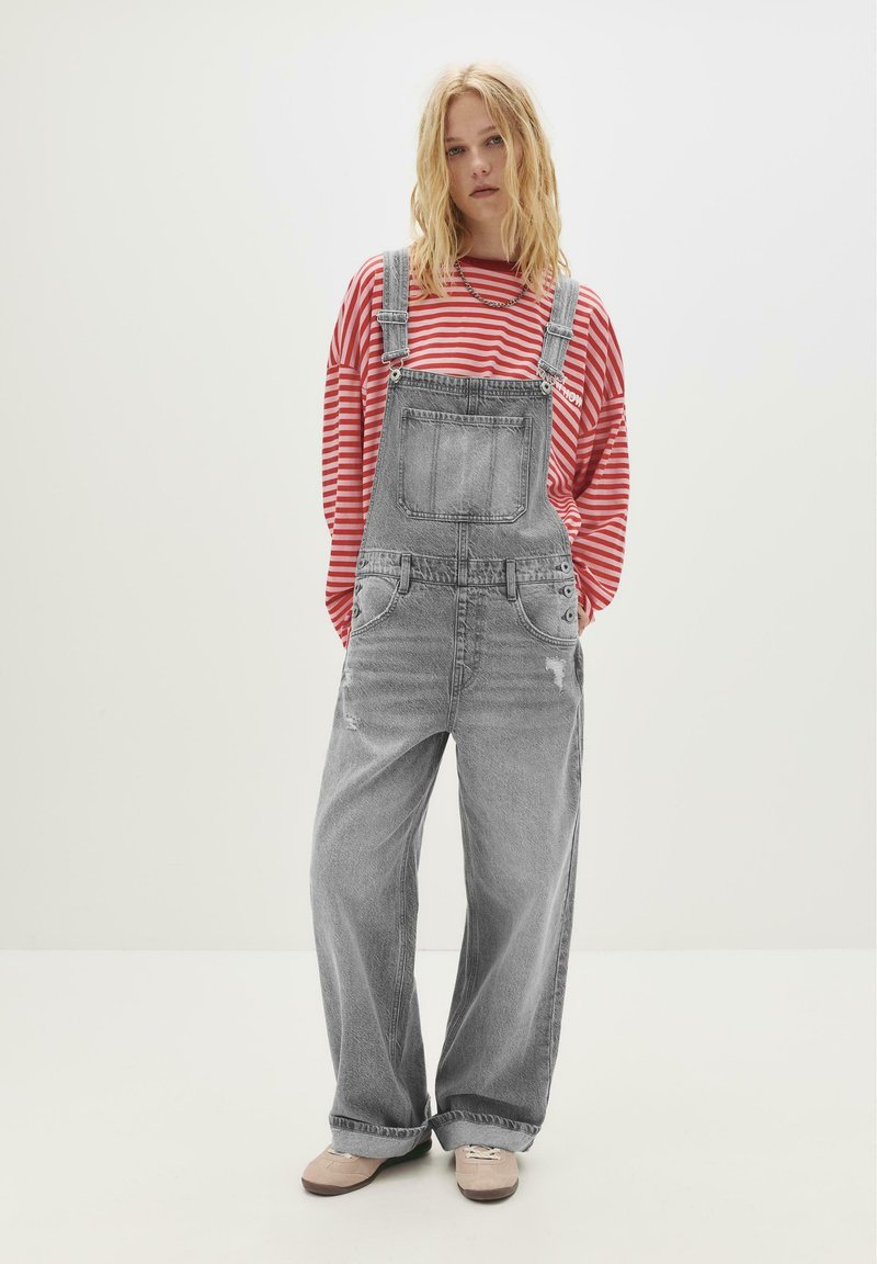Eine junge Person mit lockigem blonden Haar trägt ein rot-weiß gestreiftes Langarm-Shirt unter einer lockeren grauen Jeans-Latzhose und hellen Schuhen.