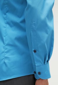 Gros plan du poignet d'une chemise bleue à manches longues avec deux boutons noirs et des coutures visibles sur le bras d'une personne.