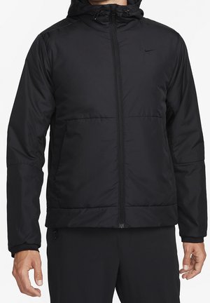 Homme portant une veste à capuche noire zippée avec un petit logo Nike sur la poitrine, debout les bras détendus le long du corps.