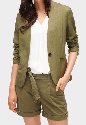 Kvinde iført olivengrøn blazer med opkærmede ærmer og matchende shorts med bælte over en hvid V-hals bluse.