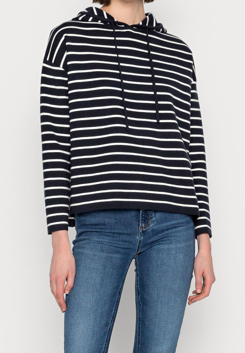 Marineblau-weiß gestreifter Hoodie, mit Kordelzug, langen Ärmeln und lockerer Passform an der Taille. Kombiniert mit blauen Jeans.