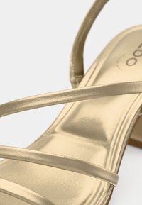 Sandale dorée à lanières avec une finition métallique, dotée de fines lanières et d'une surface lisse et brillante. Le talon est modérément élevé avec une semelle lisse.