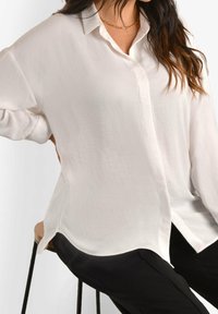 White, oversized blouse with a collared neckline and button closure. Made from a soft, textured fabric. Side slits and long sleeves.