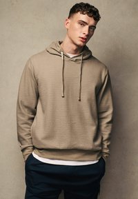 Beige strukturierter Hoodie mit verstellbaren Kordeln, Kängurutasche und gerippten Bündchen, geschichtet über einem weißen T-Shirt.