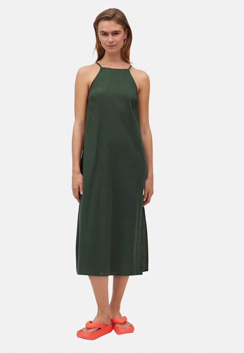 Vestido maxi de lino verde con cuello halter, aberturas laterales y textura suave, combinado con sandalias de goma naranja brillante.