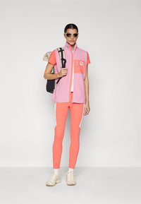 Fleece vest in pink with a chest pocket, paired with a coral t-shirt and orange leggings featuring white accents. Cream sneakers and a backpack.