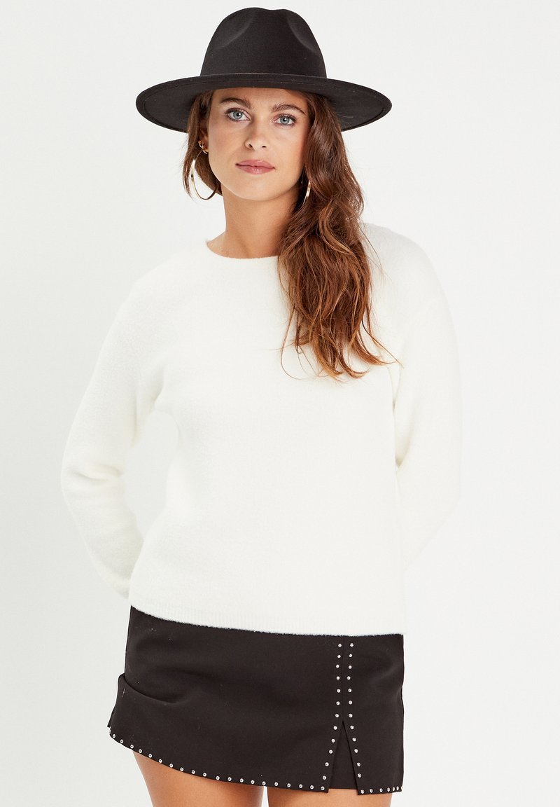 Pull crème texturé à manches longues associé à une mini-jupe noire ornée de clous en argent et d'une fente à l'avant. Chapeau en feutre noir porté.