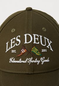 Olivengrønn baseballcaps laget av mykt stoff, med hvit brodert tekst "LES DEUX," flagg og frasen "Internasjonale sportsartikler."
